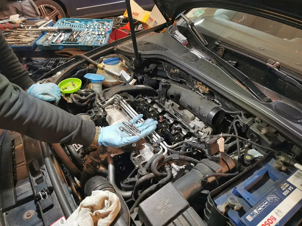 Mechanik w niebieskich rękawiczkach trzyma nowe świece żarowe, gotowe do wymiany w silniku samochodu. W tle widać skrzynkę z narzędziami.